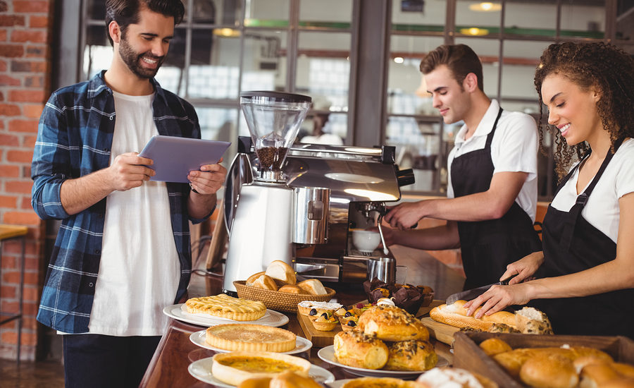 A customer using a tablet at a bakery counter with a barista preparing coffee in the background.