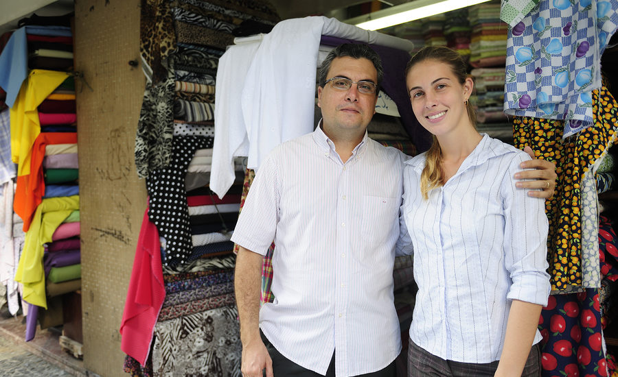 Two smiling business professionals standing in front of a fabric store with colorful rolls of fabric displayed in the background.