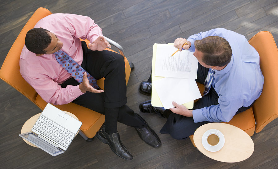 Two business professionals in a meeting, sitting across from each other on orange chairs, with a laptop, documents, and a cup of coffee on a small table nearby.