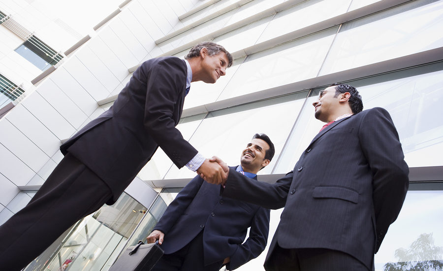 Three businessmen in suits shaking hands outside a modern glass building.