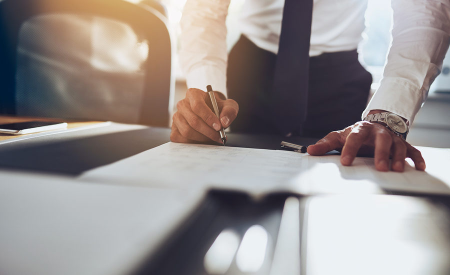 A close-up of a professional signing documents on a desk with sunlight streaming through the background.