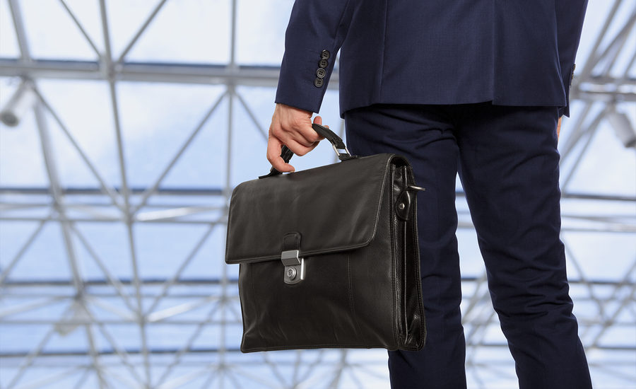 A professional carrying a sleek black leather briefcase while wearing a navy blue suit, walking through a modern building with a glass ceiling.