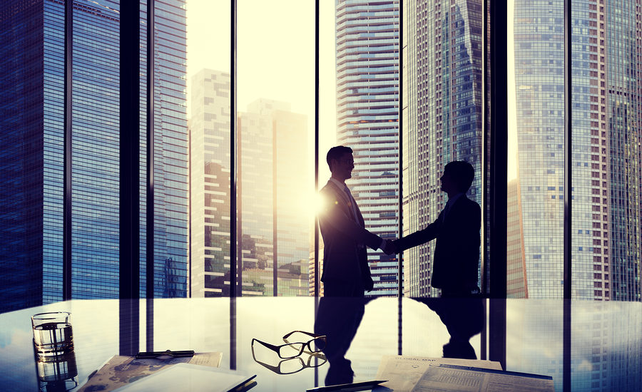 Two business professionals shaking hands in a modern office with a city skyline visible through large glass windows.