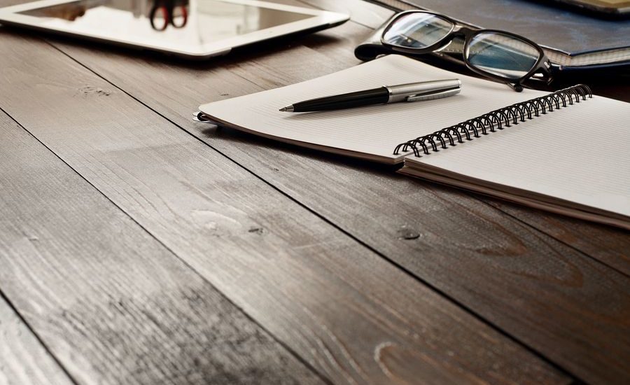 A wooden desk with an open notebook, a pen, a pair of glasses, and a tablet, creating a workspace atmosphere.