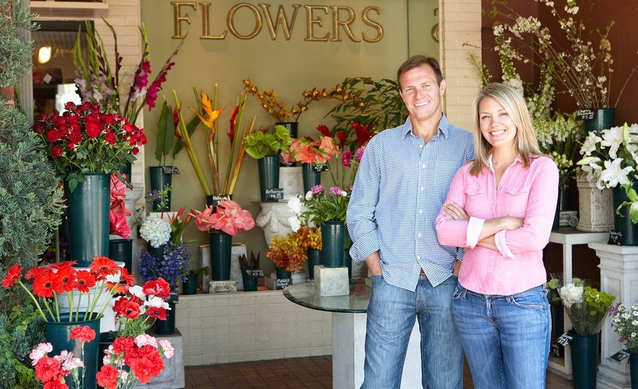Two smiling florists standing in front of their flower shop with colorful floral arrangements displayed in the background.