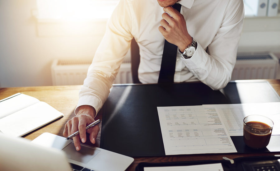 Businessman in a white shirt and tie working at a desk with financial documents and a laptop.