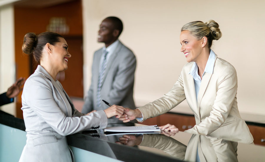 Two professional women shaking hands at a reception desk while a man in a suit stands in the background.