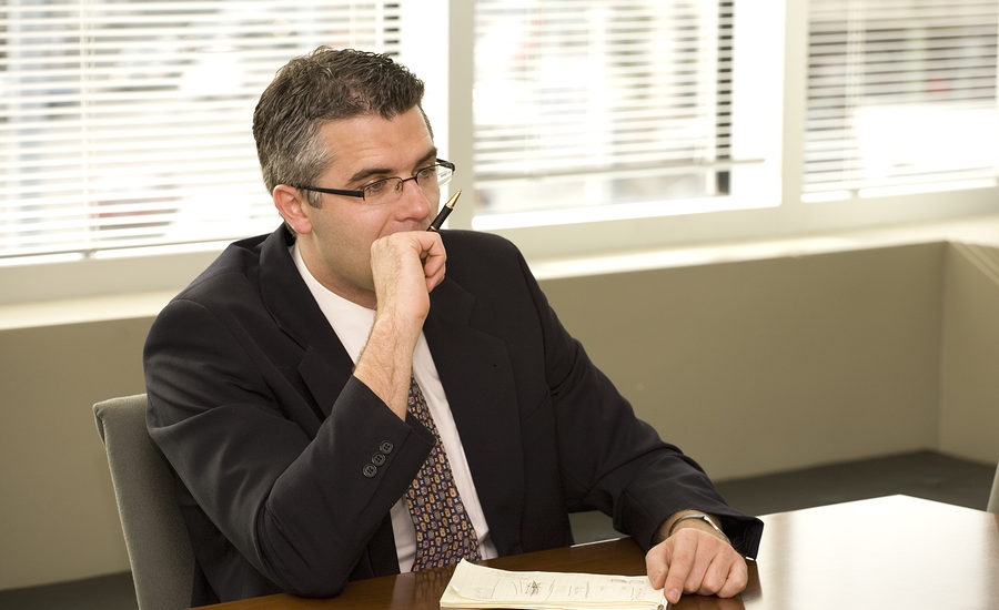 Business professional in a suit sitting at a table, holding a pen and deep in thought.