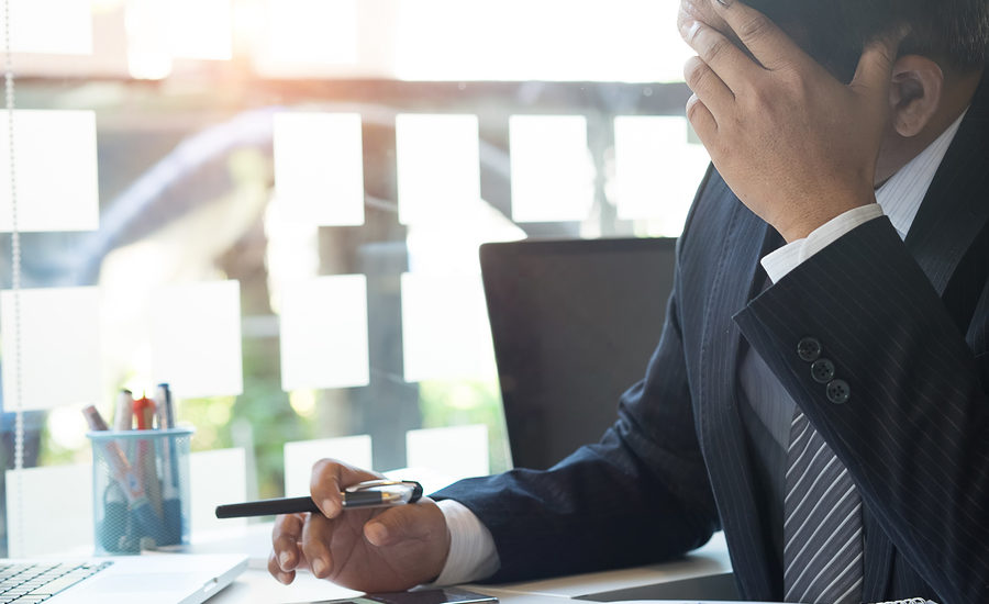 Businessman sitting at a desk, holding his head in frustration while looking at his phone.