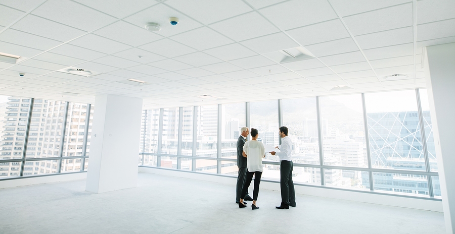 Three professionals stand in a large, empty office space with floor-to-ceiling windows overlooking a city skyline.