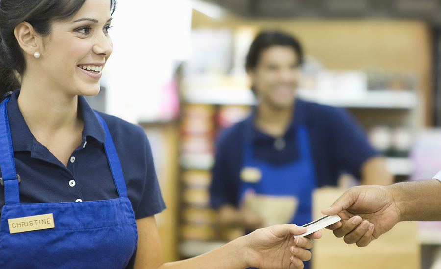 A smiling retail employee in a blue apron handing a card to a customer, with another employee blurred in the background.