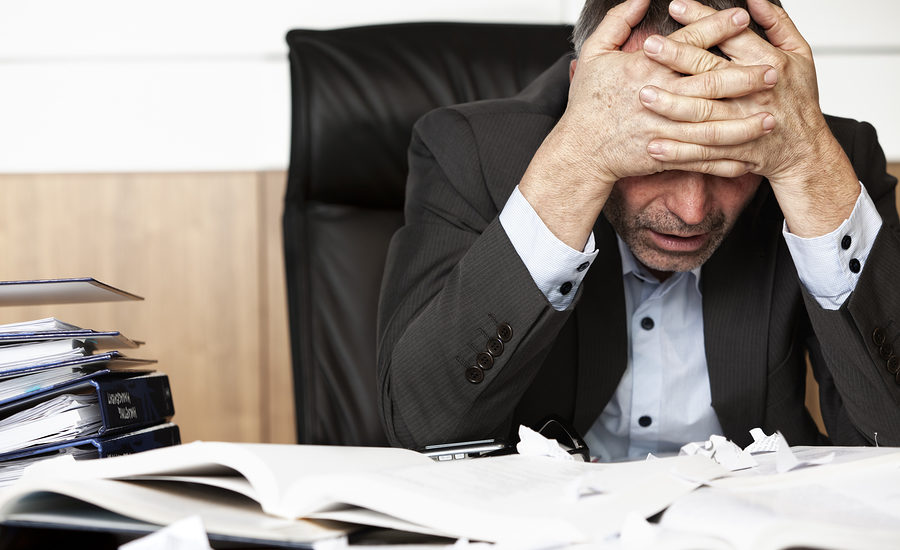 Stressed businessman sitting at his desk with head in hands and paperwork scattered around.