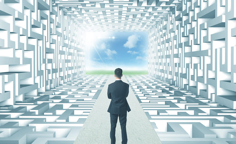 Businessman standing at the end of a maze-like tunnel, looking out towards an open, bright sky.