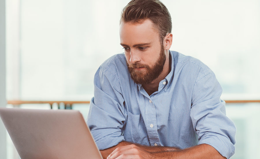 A man with a beard in a blue shirt working intently on a laptop.