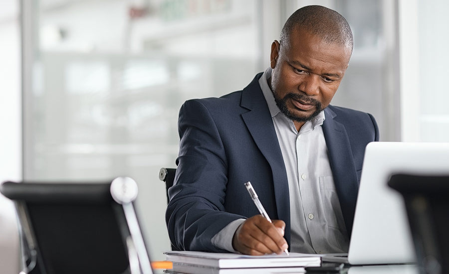A focused businessman in a suit writing notes while working on a laptop at his desk.