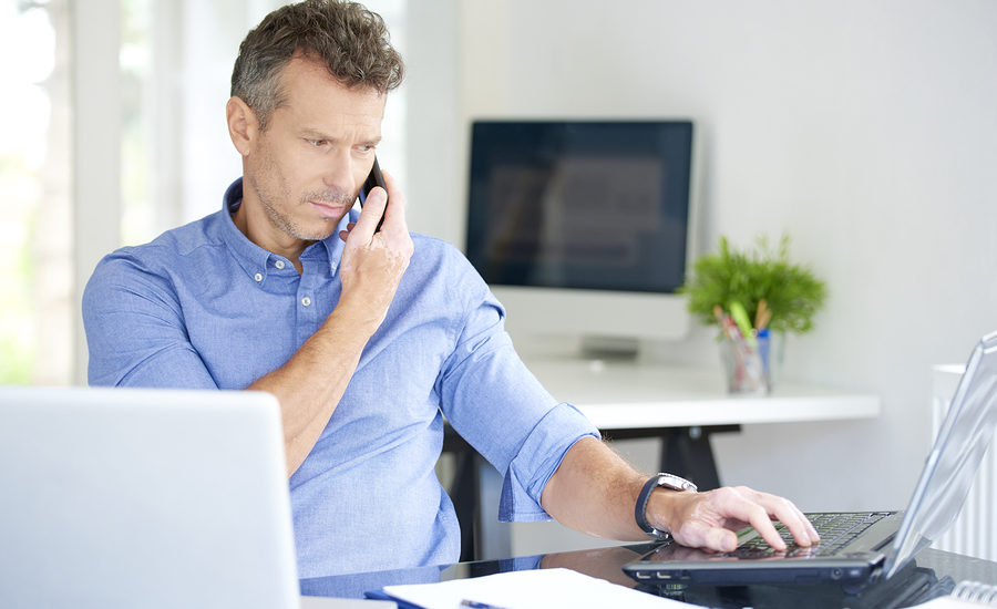 Businessman working on a laptop while speaking on a mobile phone in a modern home office.