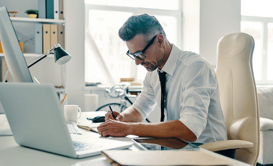 A professional man in a white shirt and tie working at his desk with a laptop and documents.
