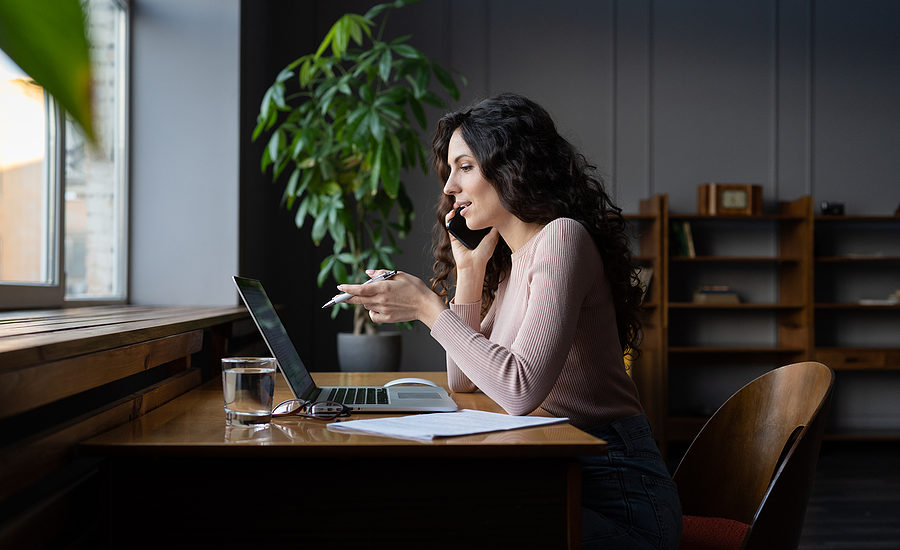 Woman talking on the phone while working on a laptop in a modern home office.