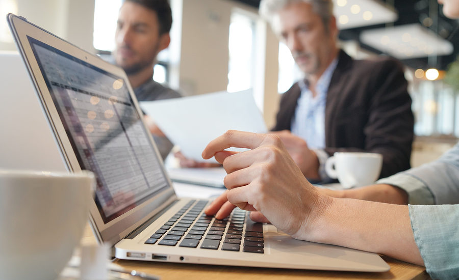 Close-up of a business professional using a laptop during a meeting, with colleagues in the background.