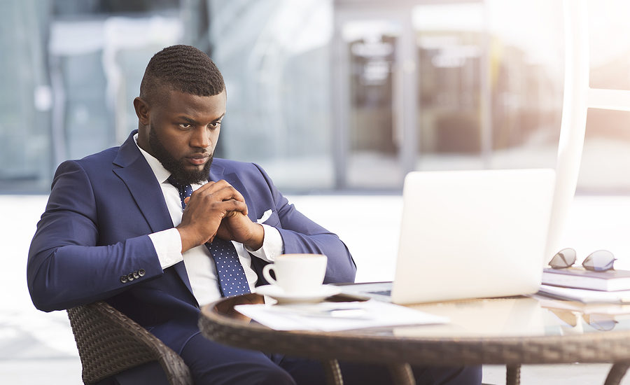 Business professional in a suit deep in thought while working on a laptop at an outdoor table.