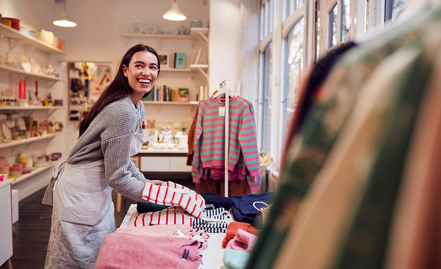 Smiling female small business owner arranging clothes in a cozy boutique with various colorful sweaters and gift items displayed on shelves.