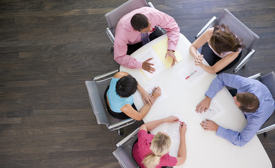 A group of five businesspeople in a meeting around a white conference table, reviewing documents and charts from a top-down perspective.