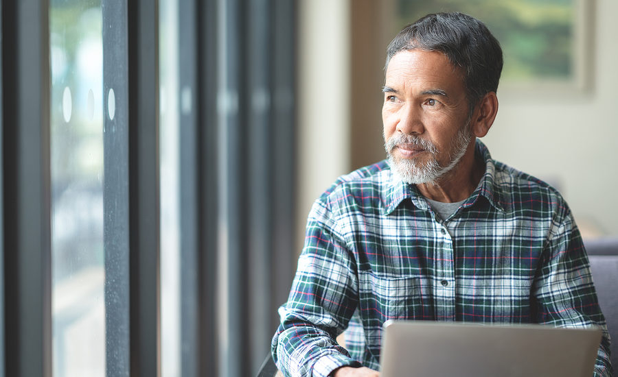 Mature man with a thoughtful expression sitting by a window with a laptop.