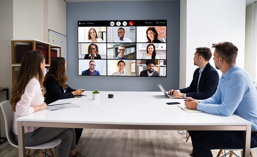 Business team in a conference room conducting a virtual meeting with colleagues displayed on a screen.