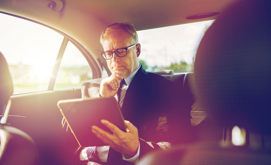 Business professional reviewing information on a tablet in the backseat of a car.
