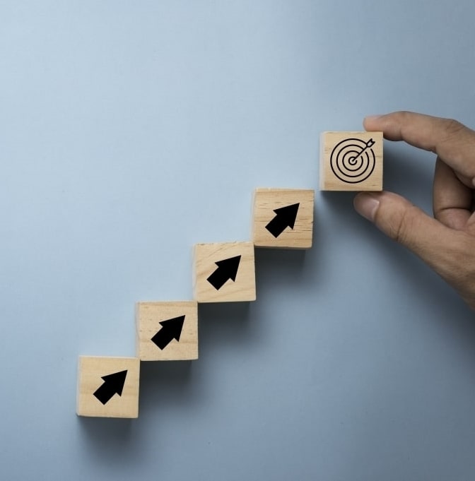 A hand placing a wooden block with a target symbol at the top of a staircase of blocks, each with an upward-pointing arrow.