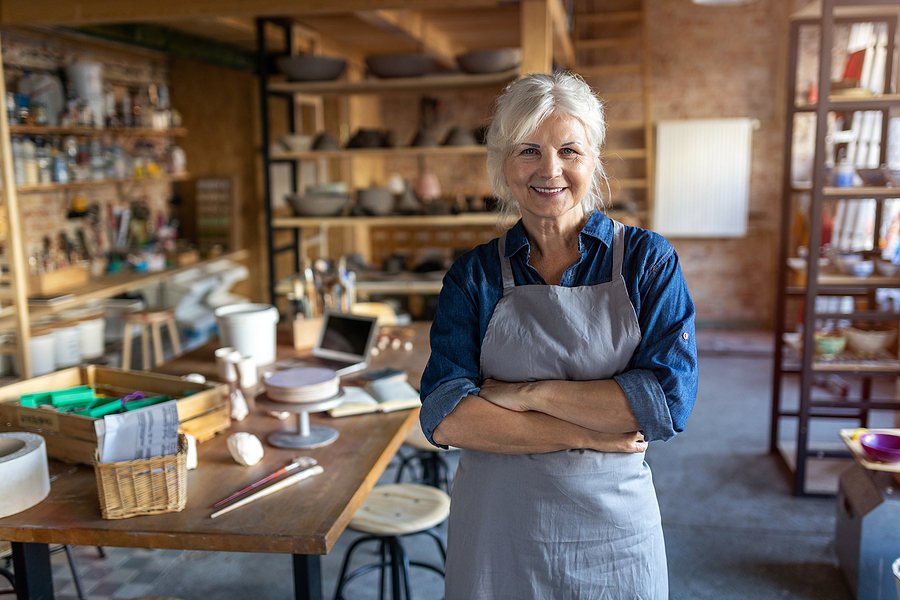 Smiling senior female artisan in a pottery studio surrounded by tools and creations.