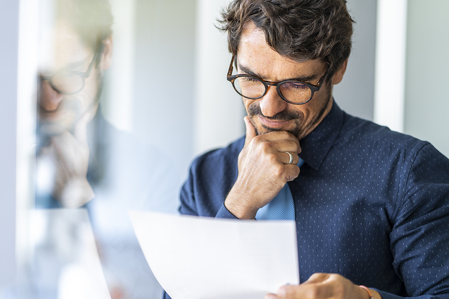 Thoughtful businessman wearing glasses, analyzing a document by a glass wall.