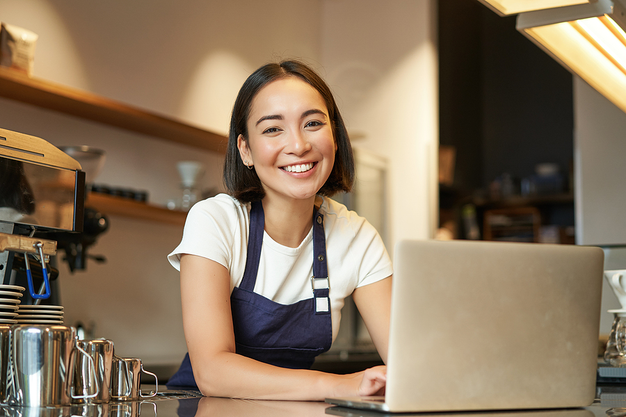 Smiling cafe owner wearing an apron, working on a laptop at the counter.