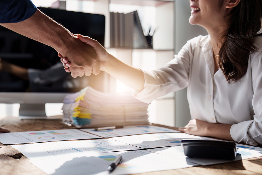 A handshake between two professionals over a desk with business documents in a bright office.