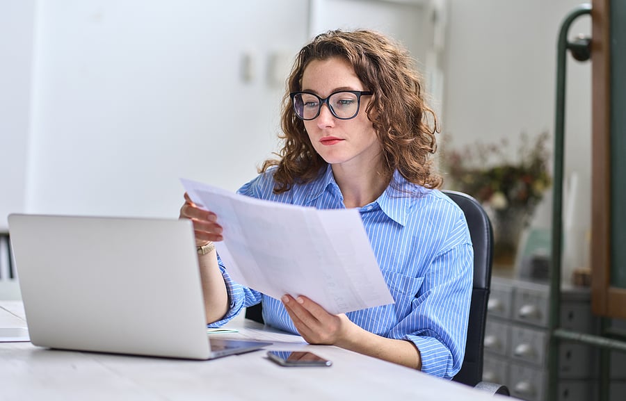 Businesswoman reviewing documents while working on a laptop at her desk in a bright office.