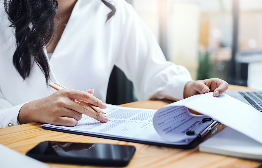 Businesswoman reviewing and marking documents on a clipboard at a desk with a smartphone.