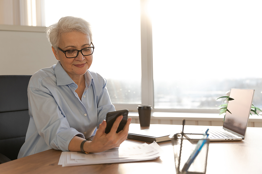 Confident businesswoman with glasses using a smartphone at her desk in a bright office.