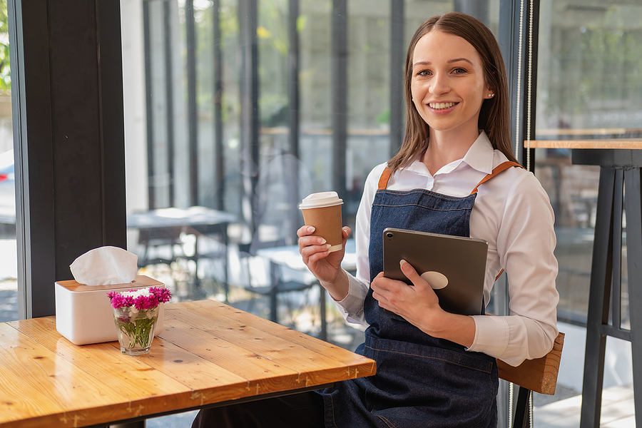 Smiling cafe owner in an apron holding a coffee cup and a tablet at a wooden table by a window.