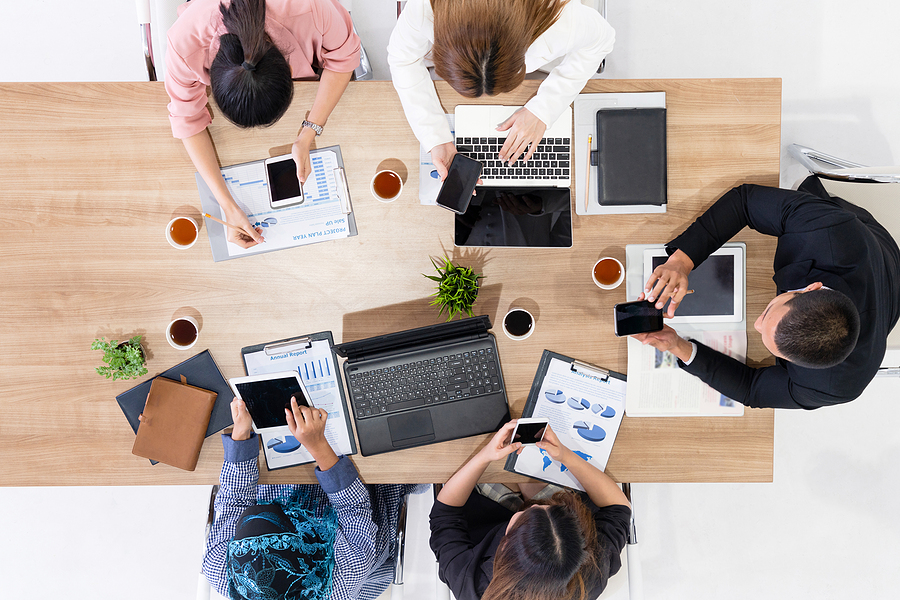 Top view of business professionals collaborating around a table with laptops, tablets, smartphones, and financial reports.
