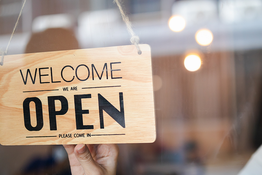Wooden "Welcome, We Are Open" sign hanging on a glass door of a shop.