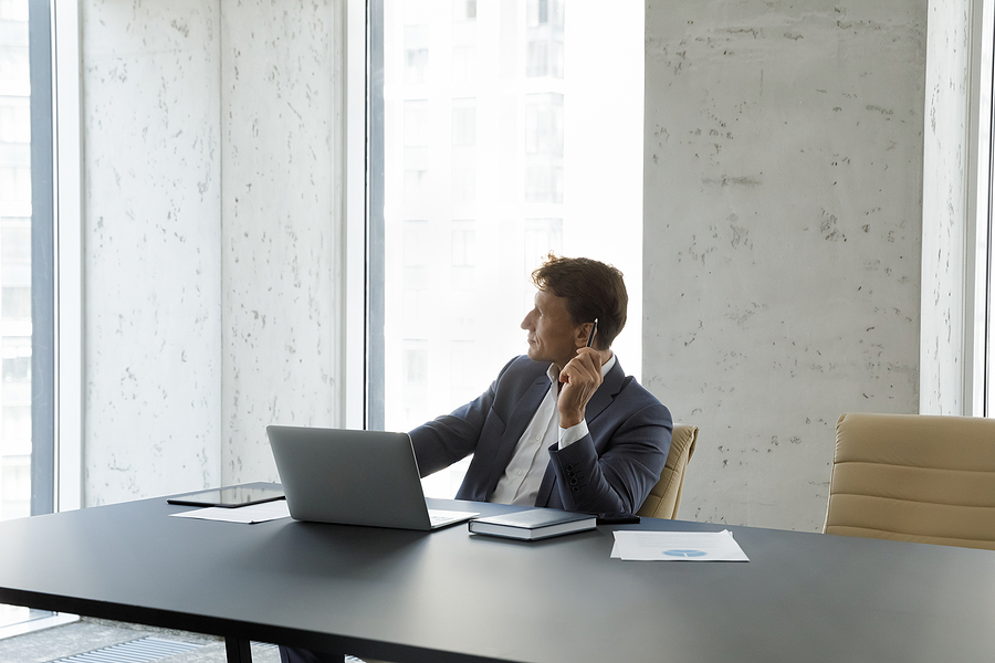 Businessman in a suit sitting at a desk with a laptop, looking thoughtfully out the window in a modern office.