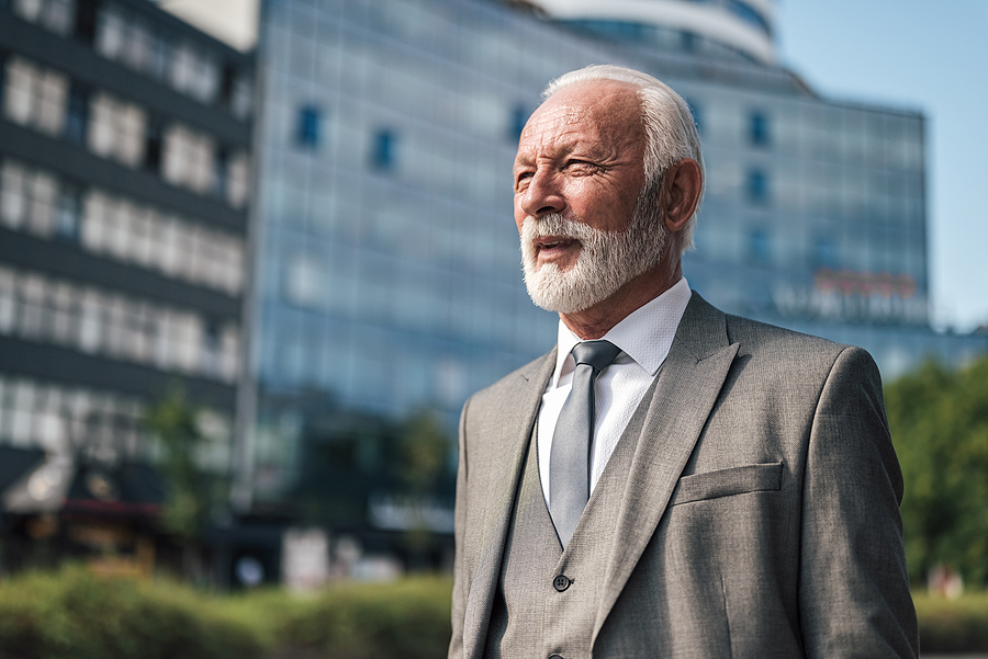 Senior businessman in a gray suit standing outdoors, looking confidently into the distance with modern office buildings in the background.