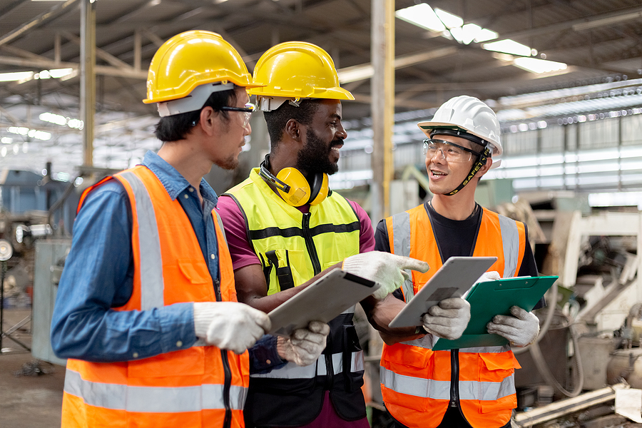 Three industrial workers wearing safety gear and helmets, collaborating and reviewing documents in a manufacturing facility.