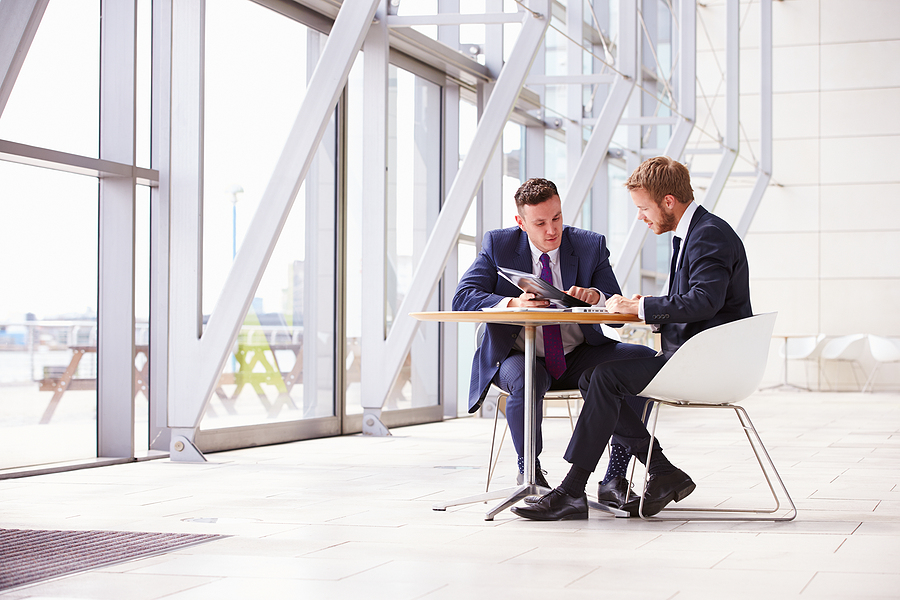 Two businessmen in suits sitting at a table in a modern office building, discussing documents.