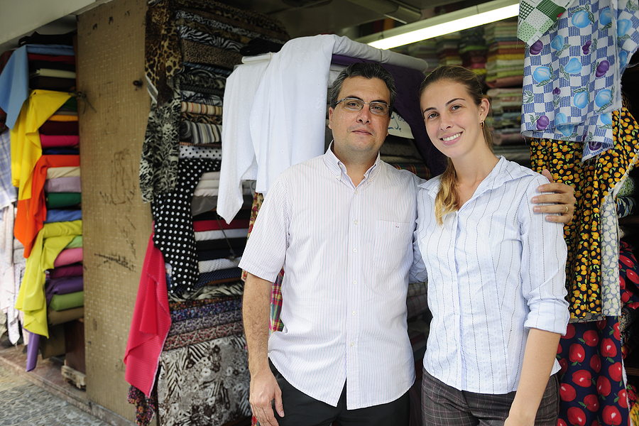 Two family business owners standing together in front of a fabric store with shelves of colorful fabrics.