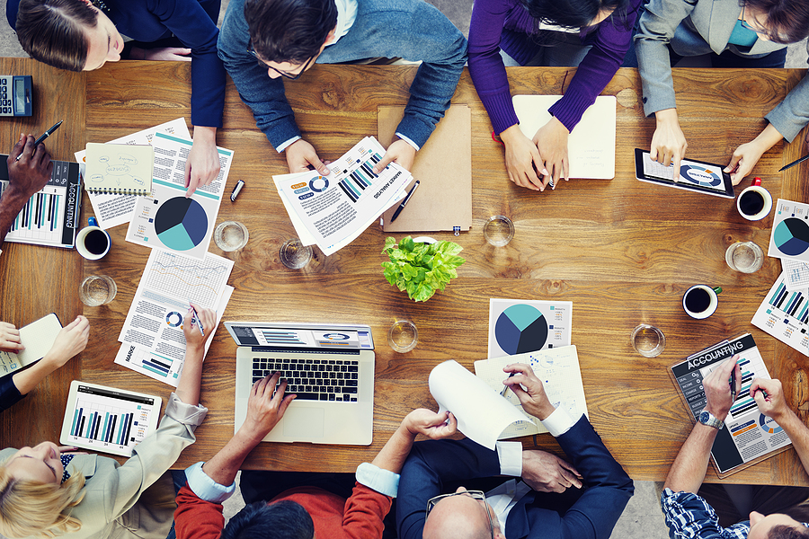 Overhead view of a diverse team analyzing data and charts around a large table with laptops, tablets, and reports.