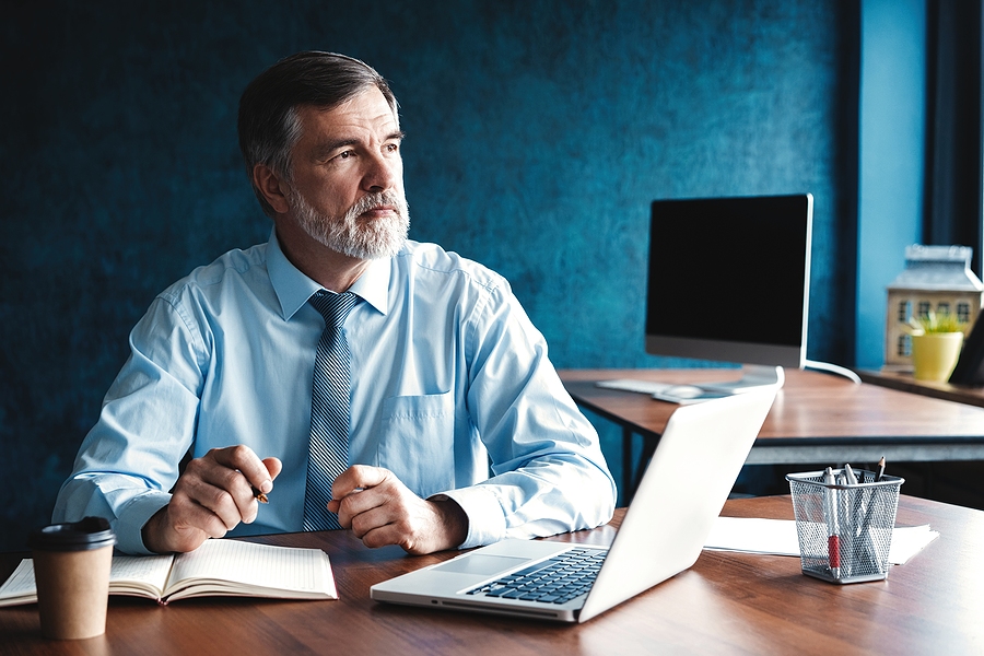 Mature businessman sitting at a desk with a laptop and notebook, appearing deep in thought.