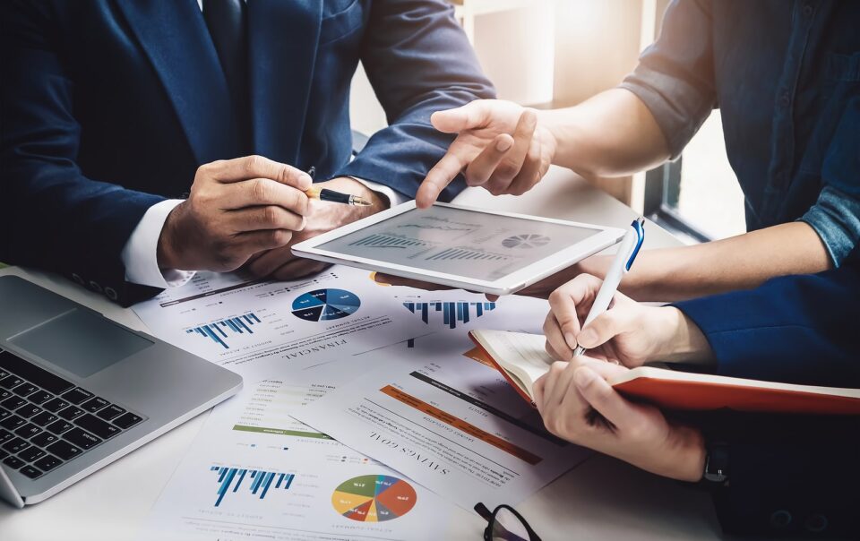 A business meeting with three people discussing financial charts and graphs. One person is holding a tablet displaying data, while another is writing notes in a notebook. A laptop and various financial documents with charts and graphs are spread out on the table.