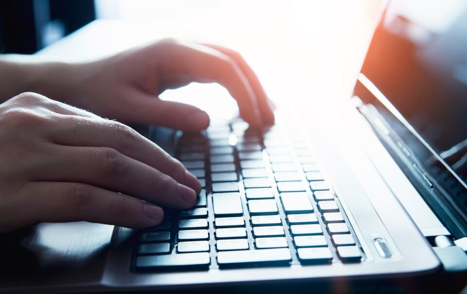 Close-up of hands typing on a laptop keyboard with sunlight reflecting off the screen.