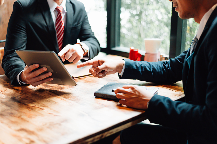 Two businessmen sitting at a wooden table, discussing and pointing at a tablet screen during a meeting